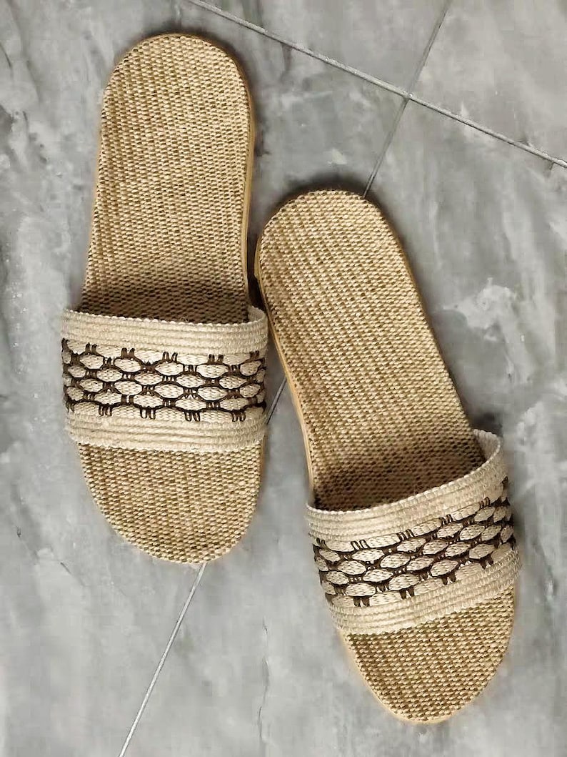 May include: A pair of beige woven sandals. The sandals have a textured, open-toe design with a decorative band featuring a repeating pattern in a darker brown color. The soles appear to be made of a light-colored wood or similar material.