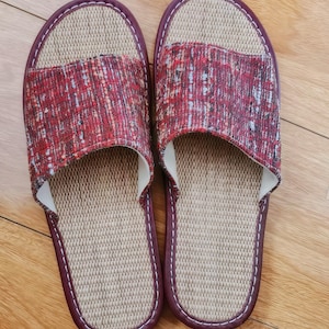 May include: A pair of open-toe slippers featuring a textured red, black, and white woven fabric upper. The footbed is made of a light beige woven material, and the edges are finished with a dark maroon trim and white stitching throughout.