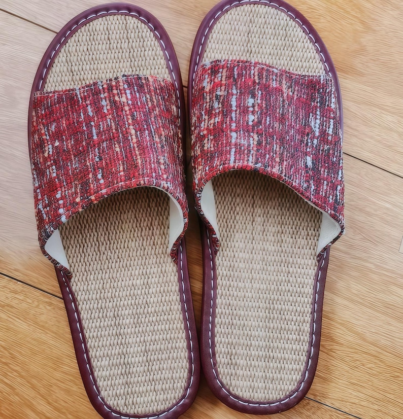May include: A pair of open-toe slippers featuring a textured red, black, and white woven fabric upper. The footbed is made of a light beige woven material, and the edges are finished with a dark maroon trim and white stitching throughout.