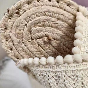 Może przedstawiać: Zbliżenie zwiniętego dywanu z juty w naturalnych odcieniach, z tkaną obw&oacute;dką. Widoczny jest spiralny wz&oacute;r dywanu, z wyraźną fakturą. Małe, białe pompony zdobią krawędź, dodając dekoracyjny element. Odpowiedni do dekoracji wnętrz.