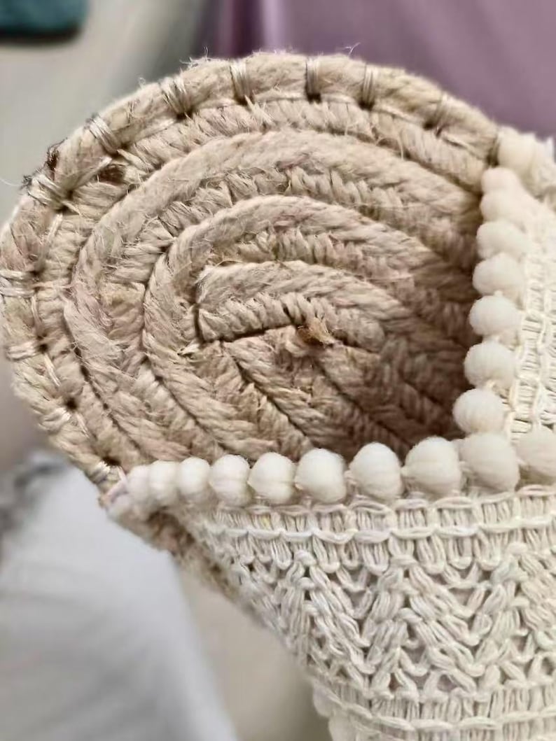 Może przedstawiać: Zbliżenie zwiniętego dywanu z juty w naturalnych odcieniach, z tkaną obw&oacute;dką. Widoczny jest spiralny wz&oacute;r dywanu, z wyraźną fakturą. Małe, białe pompony zdobią krawędź, dodając dekoracyjny element. Odpowiedni do dekoracji wnętrz.