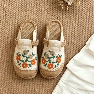 Può includere: Un paio di pantofole in tessuto beige con ricami floreali arancioni. Le pantofole hanno una suola in juta intrecciata e un cinturino intrecciato. Lo sfondo è una superficie marrone strutturata.