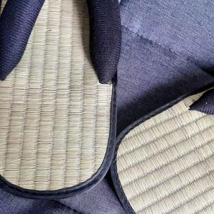 May include: Close-up of two Japanese tatami sandals, also known as zori. The sandals feature a woven straw footbed with a dark blue fabric strap. The edges are trimmed with a dark blue fabric. The sandals are resting on a blue fabric surface.