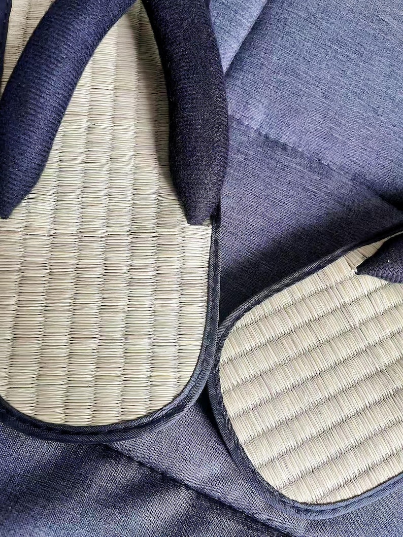 May include: Close-up of two Japanese tatami sandals, also known as zori. The sandals feature a woven straw footbed with a dark blue fabric strap. The edges are trimmed with a dark blue fabric. The sandals are resting on a blue fabric surface.