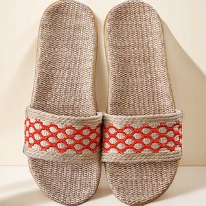 May include: A pair of beige woven slide sandals. The sandals have a red honeycomb pattern across the top strap. The soles are a light brown color. The sandals are made of a natural fiber material.