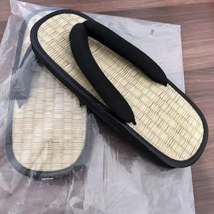 May include: A pair of traditional Japanese geta sandals. The sandals feature a woven straw footbed, a black rubber sole, and black fabric straps. The geta are displayed on a clear plastic sheet.
