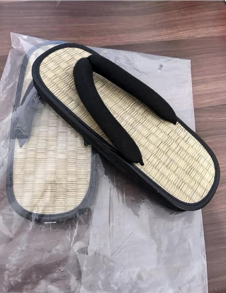May include: A pair of traditional Japanese geta sandals. The sandals feature a woven straw footbed, a black rubber sole, and black fabric straps. The geta are displayed on a clear plastic sheet.
