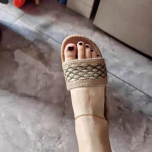 May include: A close-up view of a foot wearing a beige and brown woven slide sandal. The sandal has a decorative pattern on the strap. The toenails are painted black, and a gold anklet is visible.