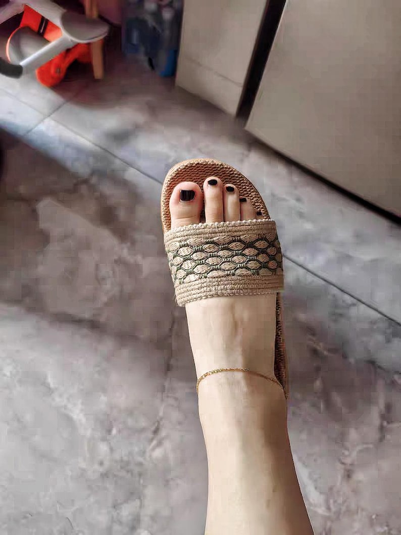 May include: A close-up view of a foot wearing a beige and brown woven slide sandal. The sandal has a decorative pattern on the strap. The toenails are painted black, and a gold anklet is visible.