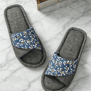 May include: A pair of gray fabric slippers with a floral design on the top strap. The strap features a blue background with white and brown flowers. The slippers have a textured appearance and a neutral color palette.