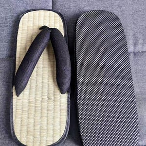 May include: A pair of traditional Japanese geta sandals. One sandal features a woven straw footbed with black straps, while the other displays a black rubber sole with a textured pattern. The sandals are designed for outdoor wear.