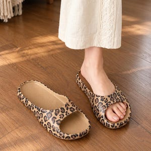 May include: A pair of slip-on sandals featuring a brown and black leopard print pattern. The sandals have a thick, rounded sole with a textured footbed and an open-toe design. One sandal is worn on a foot, while the other rests on the floor.