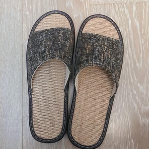 May include: A pair of open-toe slippers featuring a textured dark gray and black fabric upper strap. The footbed is made of a light beige woven material. The slippers have a dark brown trim with white stitching along the edges of the sole.
