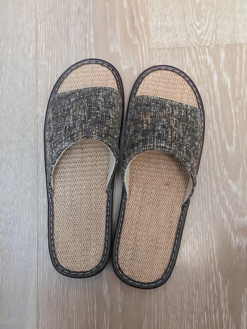 May include: A pair of open-toe slippers featuring a textured dark gray and black fabric upper strap. The footbed is made of a light beige woven material. The slippers have a dark brown trim with white stitching along the edges of the sole.
