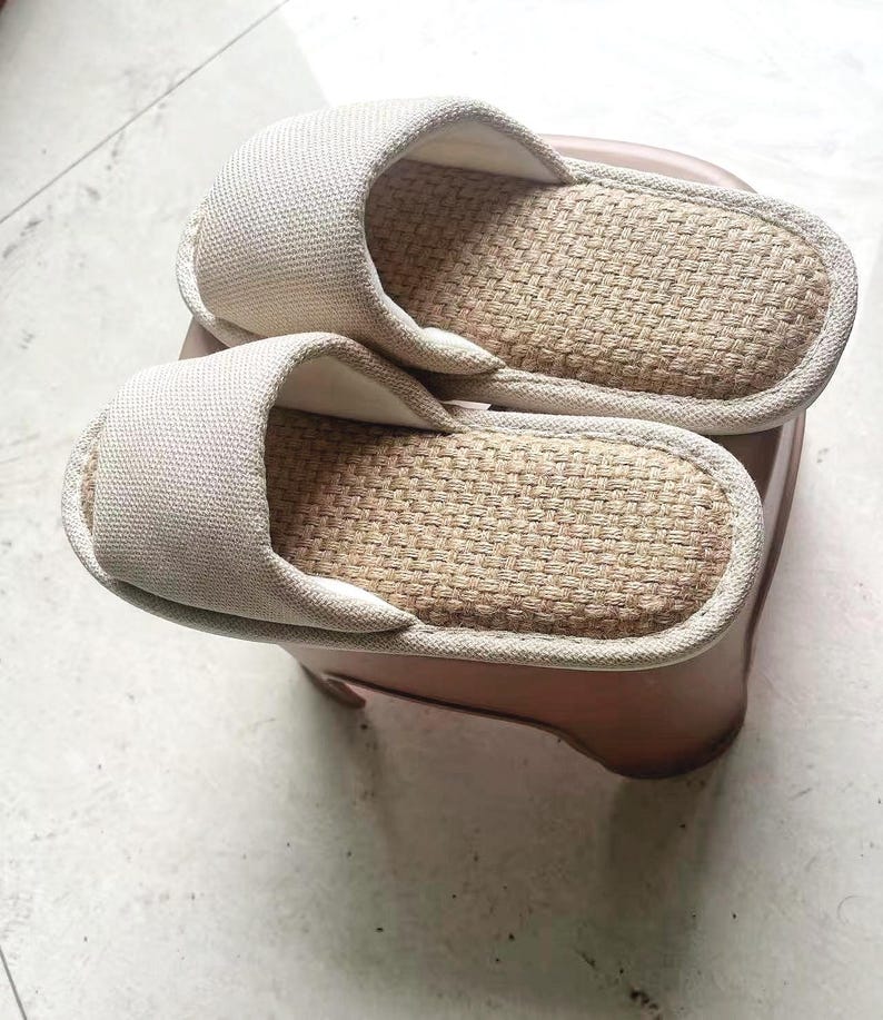 May include: A pair of beige open-toe slippers. The upper part of the slipper is made of a light beige fabric, while the footbed has a woven, natural fiber texture. The slippers are resting on a small, brown stool.