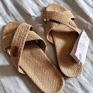 Op de afbeelding: Een paar geweven sandalen met gekruiste bandjes in een natuurlijke bruine kleur. De sandalen hebben een textuur en een kleine bruine leren patch op de band. Een blanco label is aan één sandaal bevestigd.