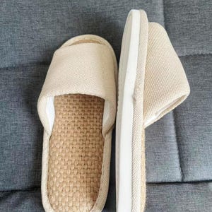May include: A pair of beige open-toe slippers. The top of the slipper is made of a woven fabric, and the sole is a light beige color with a white border. The slippers are designed for indoor use and provide comfort and relaxation.