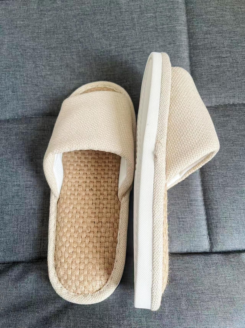 May include: A pair of beige open-toe slippers. The top of the slipper is made of a woven fabric, and the sole is a light beige color with a white border. The slippers are designed for indoor use and provide comfort and relaxation.