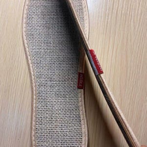 May include: Close-up of a beige woven insole with a stitched border. A red tag with the word "INSOLE" is visible. The insole is designed to fit the shape of a foot.