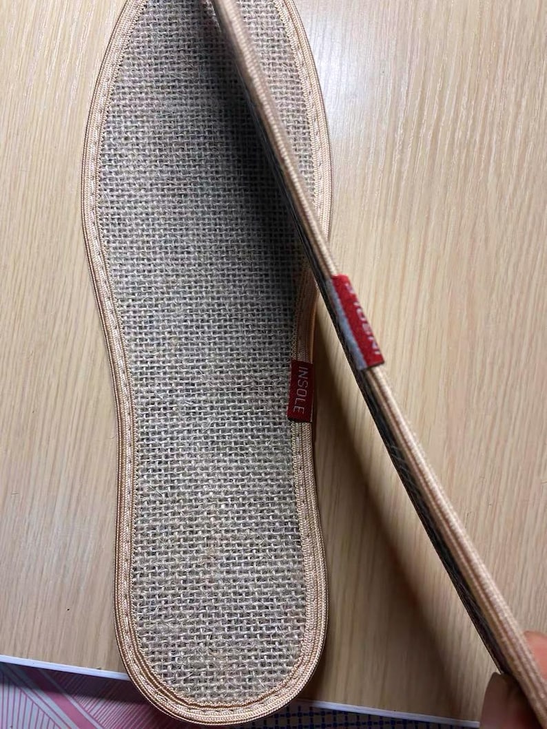 May include: Close-up of a beige woven insole with a stitched border. A red tag with the word "INSOLE" is visible. The insole is designed to fit the shape of a foot.