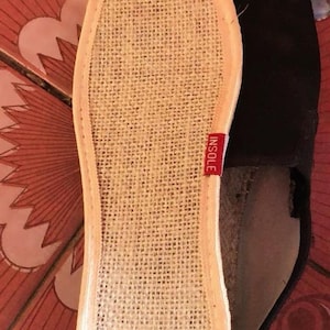 May include: Close-up of the insole of a black espadrille shoe. The insole is made of woven beige material with a red tag that reads "INSOLE". The shoe's sole is visible, and the shoe is on a patterned surface.