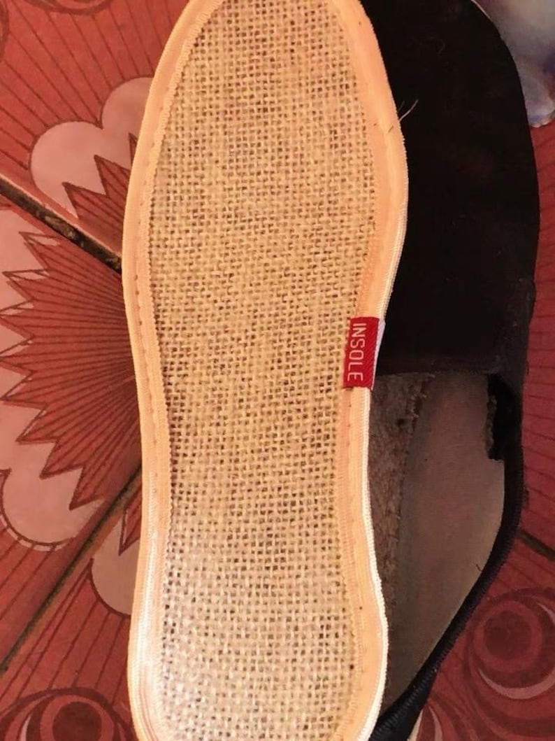 May include: Close-up of the insole of a black espadrille shoe. The insole is made of woven beige material with a red tag that reads "INSOLE". The shoe's sole is visible, and the shoe is on a patterned surface.