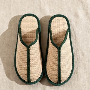 May include: A pair of beige woven slippers with a dark green trim. The slippers have a simple, open-toe design and are made of a natural fibre material. The green trim outlines the edges and the opening of the slipper.