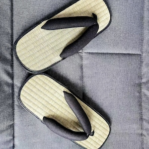 May include: A pair of traditional Japanese geta sandals. The sandals feature a natural woven straw footbed with a black fabric thong and a black border. The sandals are resting on a gray surface.