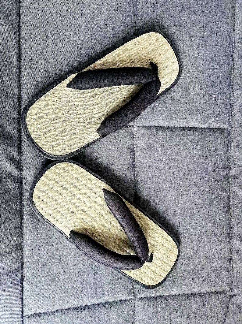 May include: A pair of traditional Japanese geta sandals. The sandals feature a natural woven straw footbed with a black fabric thong and a black border. The sandals are resting on a gray surface.