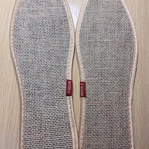 May include: Two beige woven insoles with a stitched border. Each insole has a red tag that reads "INSOLE". The insoles are designed to fit the shape of a foot and are made of a natural fiber material.