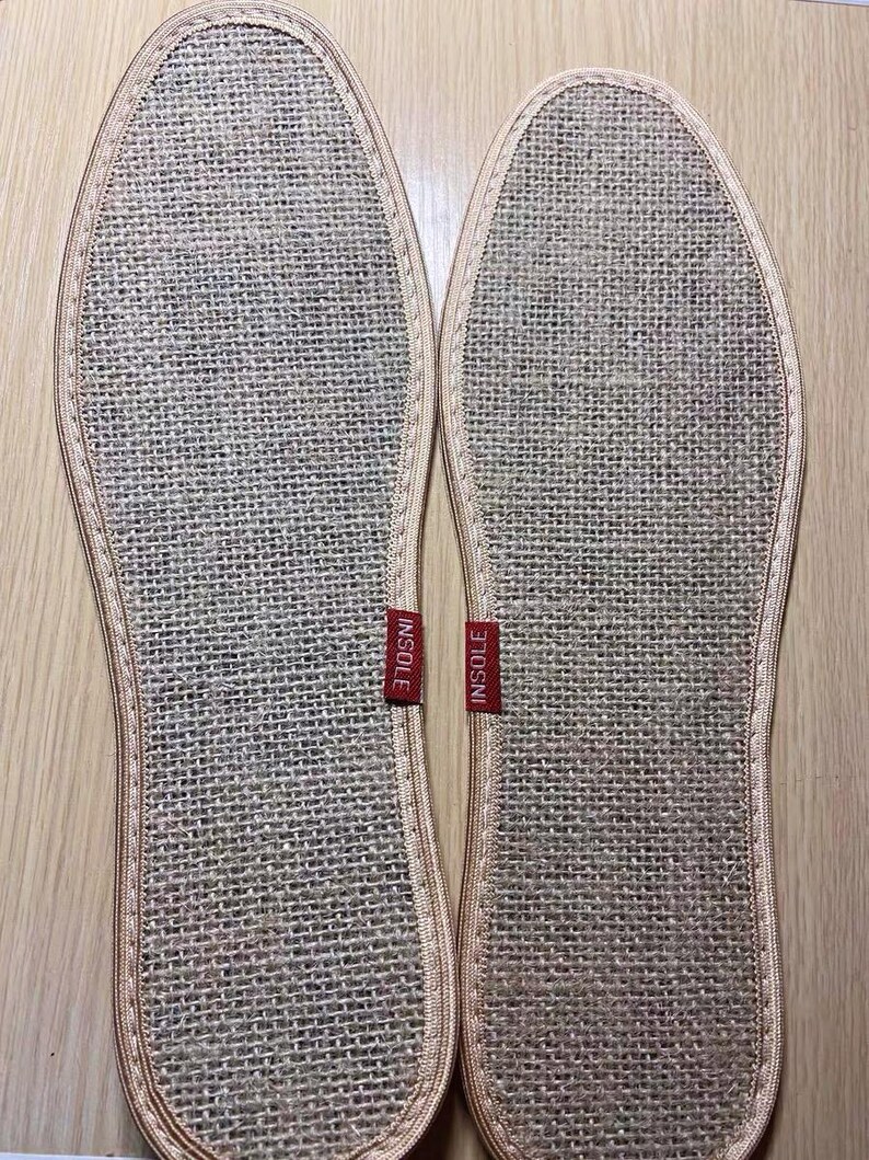 May include: Two beige woven insoles with a stitched border. Each insole has a red tag that reads "INSOLE". The insoles are designed to fit the shape of a foot and are made of a natural fiber material.