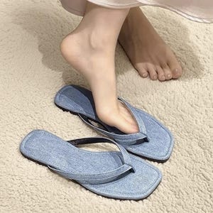 May include: Light blue denim flip-flops with black straps. The square-toed sandals are placed on a beige carpet. The flip-flops are a casual footwear option.
