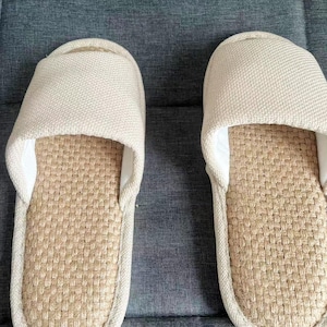 May include: A pair of beige slip-on slippers. The top of the slipper is made of a woven fabric, and the footbed has a woven texture. The slippers are designed for indoor use and provide comfort and relaxation.
