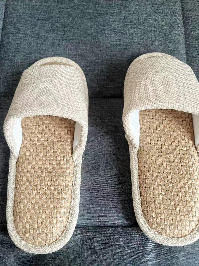 May include: A pair of beige slip-on slippers. The top of the slipper is made of a woven fabric, and the footbed has a woven texture. The slippers are designed for indoor use and provide comfort and relaxation.