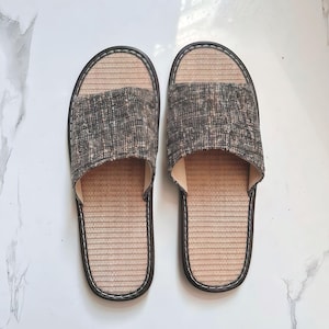 May include: A pair of open-toe slippers featuring a textured, dark gray and brown mottled fabric upper strap. The footbed is constructed from a light tan, woven material with a dark brown trim border and white contrast stitching around the edges.