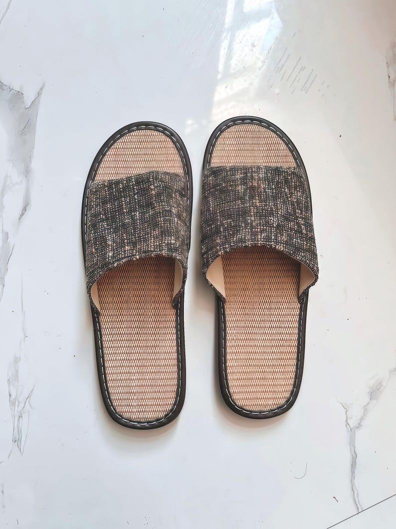 May include: A pair of open-toe slippers featuring a textured, dark gray and brown mottled fabric upper strap. The footbed is constructed from a light tan, woven material with a dark brown trim border and white contrast stitching around the edges.
