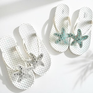 May include: White polka dot flip-flops with starfish embellishments. The flip-flops have a white thong strap and are decorated with a silver and turquoise starfish design. The flip-flops are a summer accessory.