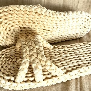 May include: A pair of natural woven sandals. The sandals are made of a light beige material, with a braided design on the footbed and straps. The sandals are shown on a neutral-colored fabric background.