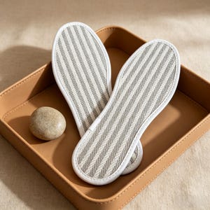 May include: Two gray and white striped insoles with white trim rest in a tan leather tray, next to a smooth, round stone. The insoles have a textured, woven appearance. The tray sits on a beige surface.