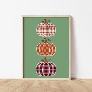 Puede incluir: Una impresión enmarcada con tres calabazas con patrones de cuadros en tonos naranja, rojo y blanco, sobre un fondo verde. Las calabazas tienen tallos marrones y están dispuestas verticalmente.