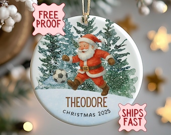 Op de afbeelding: Een ronde kerstversiering met de kerstman die voetbal speelt in een besneeuwd bos. De versiering heeft de naam "THEODORE" en het jaar "CHRISTMAS 2025" erop gedrukt. Het hangt aan een gouden draad.