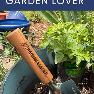 May include: A gardening trowel with a wooden handle engraved with "Hannah's Garden" rests in a teal pot filled with soil. The image includes text that reads "The Perfect Gift for Your Favorite Garden Lover."