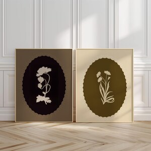 May include: Two framed botanical art prints. One features a white flower silhouette on a dark brown oval, the other a white floral design on a light brown oval. Both are set against a neutral background.