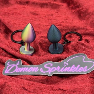 Puede incluir: Dos tapones anales con llaveros. Uno es de color arcoíris, el otro es azul oscuro. Debajo de ellos hay una pegatina con el texto "Demon Sprinkles" en escritura rosa.
