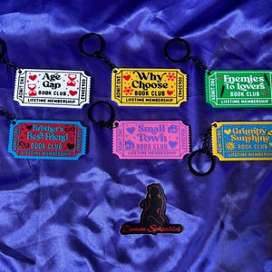 May include: A collection of colorful keychains with black key rings. Each keychain is designed as a ticket with phrases like "Age Gap Book Club" and "Enemies to Lovers Book Club." A black silhouette of a figure with the text "Demon Sprinkles" is also present.