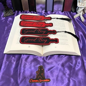 Puede incluir: Cuatro marcadores negros y rojos con las palabras "Good Boy" en escritura roja están colocados sobre las páginas abiertas de un libro. Cada marcador tiene forma de paleta con una borla. Los marcadores están sobre una superficie de satén púrpura.