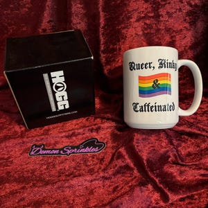 May include: A white ceramic mug with a black handle and the text "But First, Coffee" in a black, bold font. The mug is on a white surface. The mug is approximately 4 inches tall.