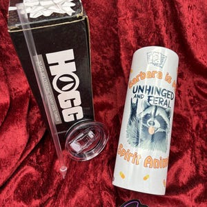 May include: A white tumbler with a raccoon graphic and the text "Barbara is UNHINGED AND FERAL Spirit Animal." Includes a lid, straw, and a black box that reads "HOGG 20.07 HOLOGRAPHIC SKINNY EDGE TUMBLER." A sticker with the text "Demon Sprinkles" is also present.