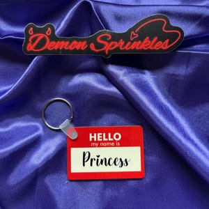 May include: Red name tag with the text "HELLO my name is Princess" on a blue satin background. A silver key ring is attached to the name tag.
