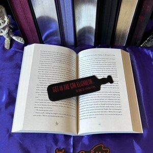 Puede incluir: Un libro abierto con un marcapáginas negro que dice "GET IN THE CAR ELIZABETH.. S3E3 IYKYK" en texto rojo. El libro está sobre una superficie púrpura con otros libros en el fondo. Una pegatina de "Demon Sprinkles" está en la parte inferior.